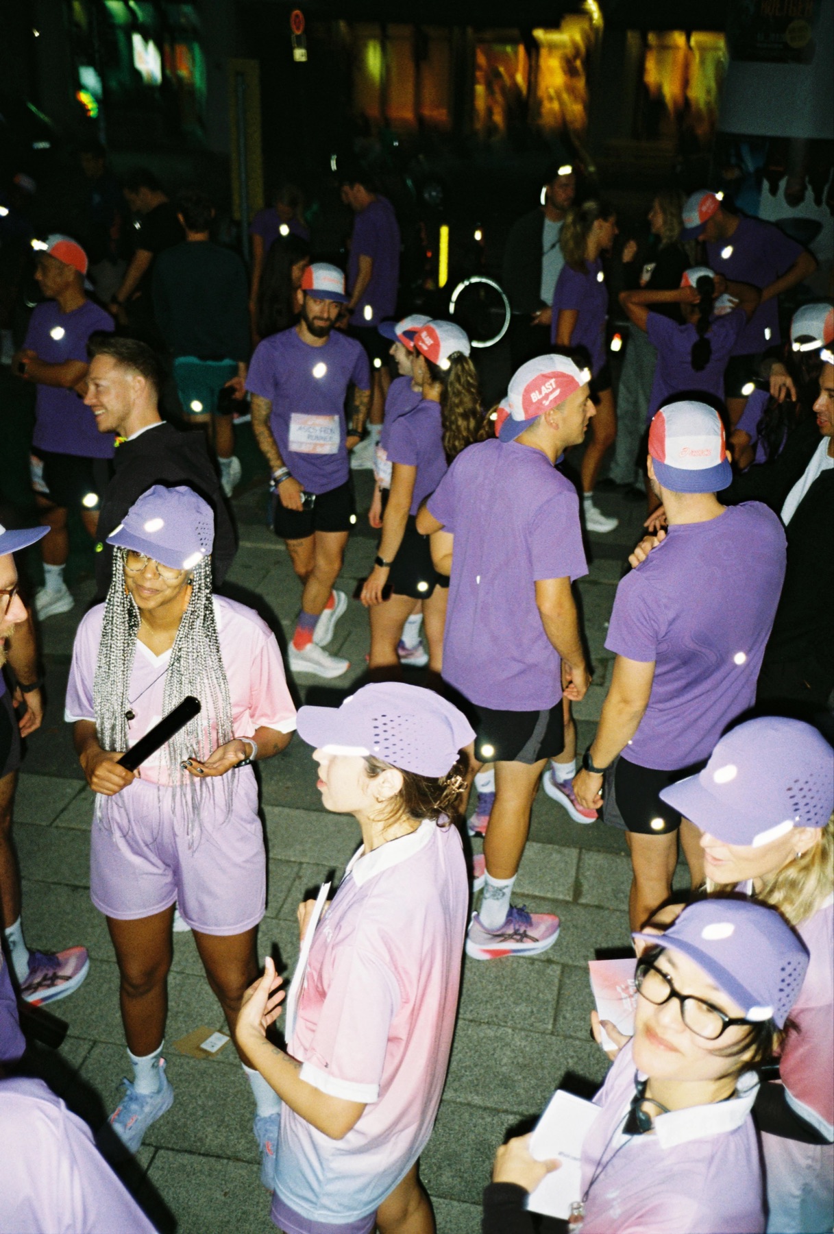ATH.Crew runners in purple shirts and caps gathered at night, lit by camera flash